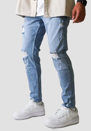 Mand iført lyseblå revnede skinny jeans og hvide sneakers står mod en ensfarvet baggrund.