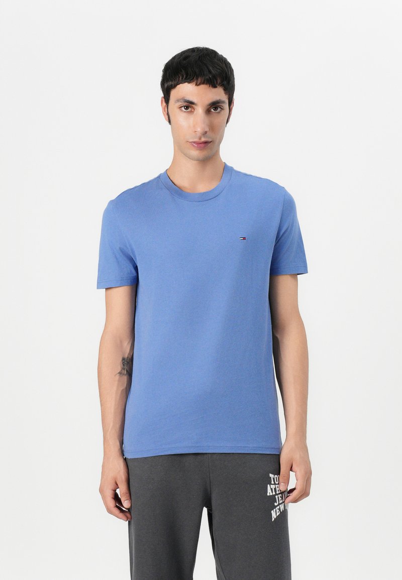 Camiseta de algodón azul con cuello redondo y mangas cortas. Presenta un pequeño logo en rojo, blanco y azul en el pecho. Textura suave.