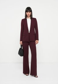 Bordeaux op maat gemaakt pak met een getailleerde blazer, brede broekspijpen en zijzakken; gecombineerd met een wit T-shirt en een zwarte handtas.