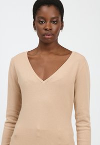 Beige Strickpullover mit V-Ausschnitt, langen Ärmeln und einer weichen Textur. Das Design besitzt eine figurbetonte Silhouette und minimale Details.