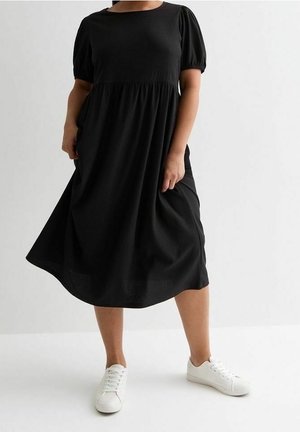 Donna con un vestito midi nero largo con maniche corte a sbuffo e sneakers bianche, in piedi davanti a uno sfondo chiaro e uniforme.