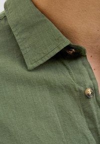 Groene linnen overhemdkraag met een gestructureerde afwerking en metalen knopen. Kenmerken zijn een subtiele weving en stikdetails langs de kraag.