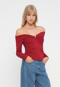 Top rojo con un hombro al descubierto, de mangas largas, con tejido texturizado, lados fruncidos y un detalle en dorado en el centro. Combinado con jeans de tiro alto azul.