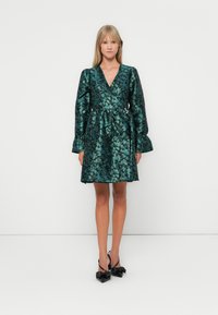 Green floral patterned dress with a V-neck, long puffed sleeves, and a cinched waist. Worn with black heeled sandals featuring bow accents.