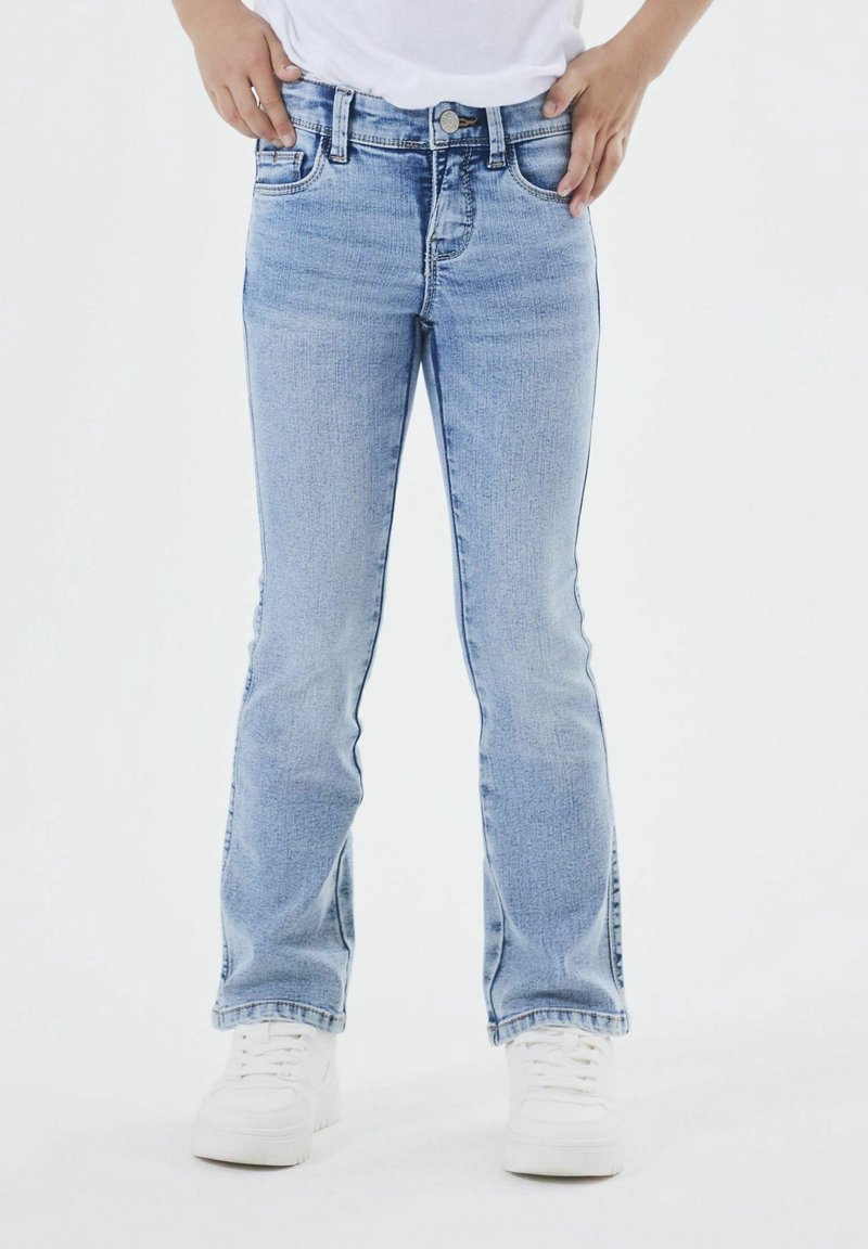 Personne portant un jean droit bleu clair et des baskets blanches, debout les mains posées sur les hanches.