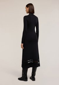 Donna in un vestito midi nero a maniche lunghe con dettagli a taglio sul bordo, che indossa stivali neri al ginocchio e orecchini dorati, rivolta di spalle.