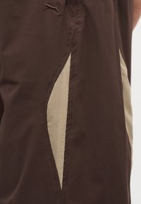 Καφέ υφασμάτινη βερμούδα με μπεζ διχτυωτή πλευρική επένδυση και κεντημένο λογότυπο Puma κοντά στη μέση.