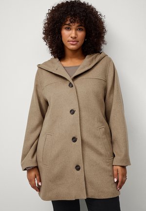 Kvinde iført en brun hættetrøje med fire knapper og sidelommer, stående mod en ensartet lys grå baggrund.