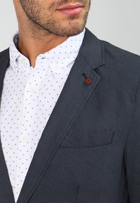 Marinefarbener Blazer mit Struktur und fallendem Revers, kombiniert mit einem weißen Hemd, das kleine marineblaue Muster und einen Knopfakzent am Blazer aufweist.