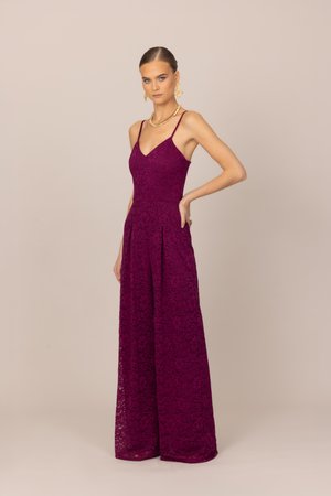 Γυναίκα όρθια με το χέρι στη μέση, φοράει αμάνικη ολόσωμη φόρμα από σκούρο μοβ δαντέλα με φαρδιά πόδια, χρυσό κολιέ και σκουλαρίκια σε μπεζ φόντο.