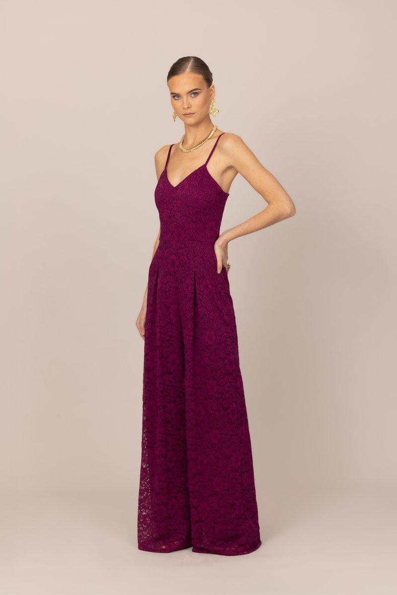 Γυναίκα όρθια με το χέρι στη μέση, φοράει αμάνικη ολόσωμη φόρμα από σκούρο μοβ δαντέλα με φαρδιά πόδια, χρυσό κολιέ και σκουλαρίκια σε μπεζ φόντο.