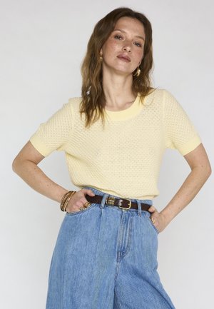 Donna con capelli castano chiaro indossa una maglia a maniche corte di colore giallo pallido, jeans blu a vita alta e gioielli dorati, in piedi con le mani sui fianchi.