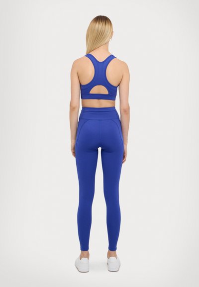Modrý sportovní crop top s výřezem na zádech, kombinovaný s modrými legíny s vysokým pasem. Hladký, elastický materiál; bílé boty.
