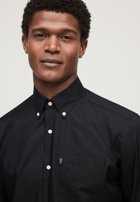 Man with short curly hair wearing a black button-up shirt with a chest pocket and a neutral expression on a light background.