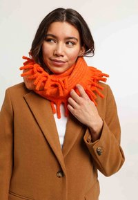 Femme portant un manteau marron et une écharpe frangée orange vif, regardant sur le côté avec une main touchant l'écharpe.