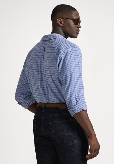 Niebieska koszula w kratę z guzikami, z podwiniętymi rękawami, noszona z ciemnymi dżinsami i brązowym paskiem. Tkanina wydaje się lekka i teksturowana.