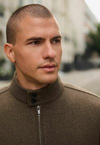 Jachetă din lână maro, cu guler înalt, prevăzută cu un fermoar și două butoane negre, cu o textură netedă și detalii de design minimaliste.