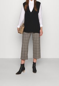 Svart stickad ärmlös topp över en vit skjorta med knappar, grå rutiga capribyxor och svarta ankelboots med blockklack.