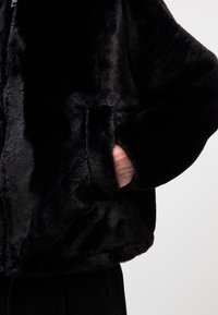 Black faux fur jacket featuring a soft texture, side pockets, and a relaxed fit, with visible stitching and a zip closure.