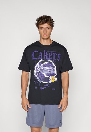 Crna oversized majica s ljubičastim natpisom "Lakers", grafikom košarke i Nike logom. Nosite je s svijetlosivim perforiranim šortsevima.