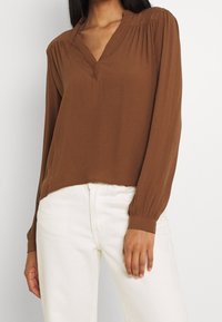 Femme portant un chemisier marron à manches longues avec un col en V et des épaules plissées, ainsi qu'un pantalon blanc taille haute, sur un fond uni.