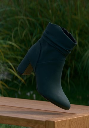 Bottine bleu foncé à talon haut carré et revers plié, présentée au-dessus d'une table en bois avec un fond de feuillage vert.