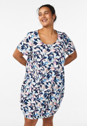 Mujer sonriente con cabello oscuro recogido en un moño, que lleva un vestido de manga corta hasta la rodilla con patrones florales en azul, rosa y blanco, de pie con las manos detrás de la espalda.