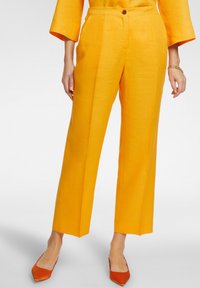 Pantaloni gialli di lino con taglio dritto, dotati di chiusura frontale con bottone e tasche laterali. Portati con ballerine arancioni a punta.
