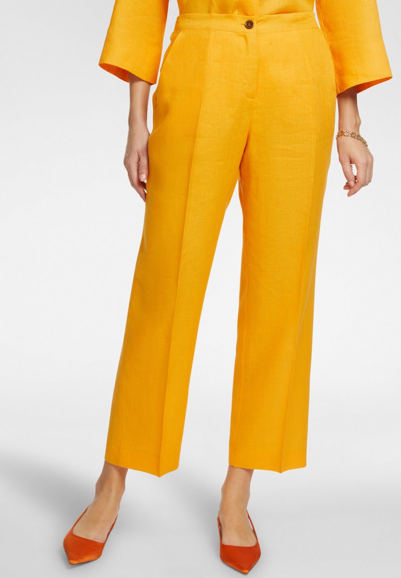 Pantaloni gialli di lino con taglio dritto, dotati di chiusura frontale con bottone e tasche laterali. Portati con ballerine arancioni a punta.
