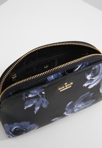 Černá kosmetická taška s modrým květinovým vzorem, zlatým zipem a logem značky. Obsahuje jednu hlavní přihrádku a vnitřní zipovou kapsu.