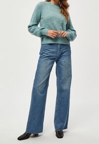 Maglione lavorato a maglia blu chiaro e peloso con maniche lunghe; abbinato a jeans blu a vita alta e gamba larga; scarpe nere con tacco completano l'outfit.