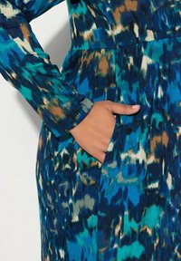 Sininen ja turkoosi kuosiset mekko, jossa on pitkät hihat, istuva vyötärö ja sivutasku, valmistettu sileästä ja kevyestä kankaasta.