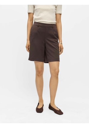 Kvinde iført mørkebrune knælange skræddersyede shorts, beige kortærmet top og mørkebrune vævede flade sko, stående mod ensfarvet baggrund.