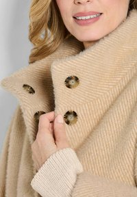 Beige visgraatjas met een hoge kraag. Het heeft grote, ronde, goudkleurige knopen en een zachte, gestructureerde stof. Gehaakte mansjetdetails zichtbaar.