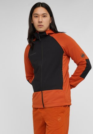 Giovane con capelli lunghi e scuri indossa una giacca arancione e nera con zip e pantaloni arancioni coordinati, in piedi con le mani dietro la schiena.