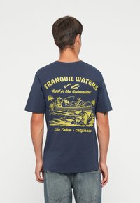 Mørkeblå bomulds t-shirt med et gult fiskerelateret grafik på ryggen med tekst: "TRANQUIL WATERS," "Reel in the Relaxation," og sted.