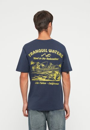 Navy blue cotton t-shirt featuring a yellow fishing-themed graphic on the back with text: "TRANQUIL WATERS," "Reel in the Relaxation," and location.