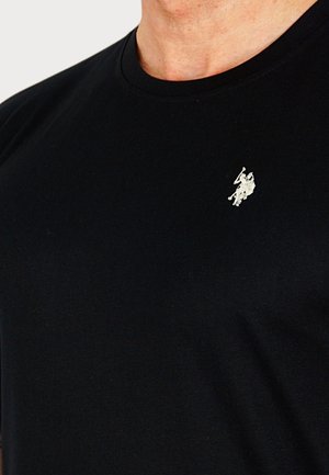 Čierne tričko s okrúhlym výstrihom, ktoré nosí osoba, s malým bielym vyšívaným logom hráča póla na ľavej strane hrudníka.