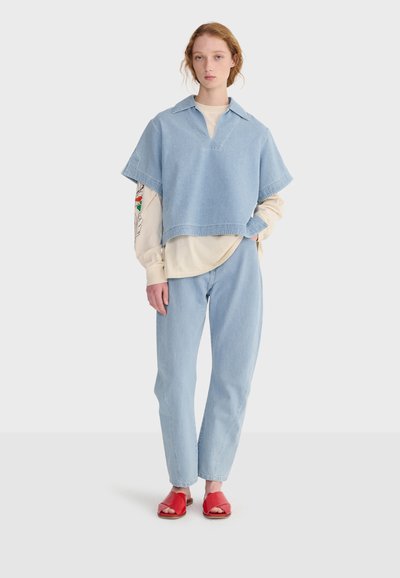 Jeune femme aux cheveux châtain clair portant un pantalon et un haut en denim bleu clair sur un chemisier beige à manches longues, assortis de sandales rouges à bout ouvert.