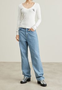 Vit långärmad T-shirt med V-ringning och broderad logotyp, parat med ljusblå vida jeans och metalliska plattformsslip-on skor. Släta texturer.