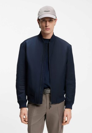 Giubbotto bomber navy con chiusura frontale a zip, polsini a costine e texture liscia; indossato su una camicia navy e abbinato a pantaloni marrone chiaro.