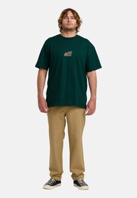 Camiseta de algodón verde oscuro de manga corta, con un gráfico de montañas rojas y naranjas en la parte frontal. Combinada con pantalones caqui y zapatillas negras.