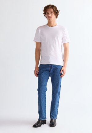 Jonge man met krullend haar, gekleed in een eenvoudig wit T-shirt, blauwe spijkerbroek en zwarte leren laarzen, staand tegen een witte achtergrond.