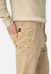 Beige corduroy pants with vertical ribbing, featuring a black logo patch on the pocket, side pockets, and a relaxed fit.