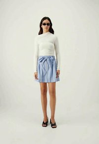 Γυναίκα με μαύρα γυαλιά ηλίου, λευκή μπλούζα με μακριά μανίκια, μπλε ριγέ φούστα με φιόγκο και μαύρες ανοιχτές γόβες που στέκεται μπροστά σε απλό φόντο.