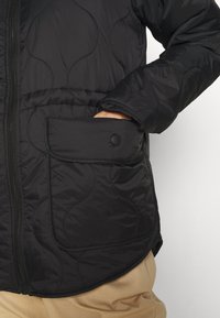 Svart vadderad jacka med zip-stängning, med en sidoficka som säkras av en svart tryckknapp och en jämn, glansig yta.