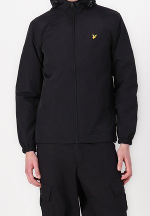 Person trägt eine schwarze Zip-up-Jacke mit elastischen Bündchen und einem gelben Vogel-Logo auf der Brust, kombiniert mit schwarzen Cargo-Hosen.