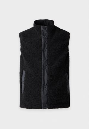 Un mannequin masculin se tient devant un fond blanc, vêtu d’une veste polaire noire zippée, d’un jean bleu ample et de baskets blanches.