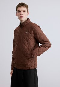 Καφέ μπουφάν καπιτονέ με ψηλό γιακά, κλείσιμο με φερμουάρ, πλαϊνές τσέπες και διακριτική λεπτομέρεια λογότυπου στο στήθος. Λεία υφή.