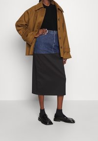 Brun ullrock, svart skjorta, denimkjol med svart nederdel och polerade svarta loafers med mörka strumpor. Platt ställning, neutral bakgrund.